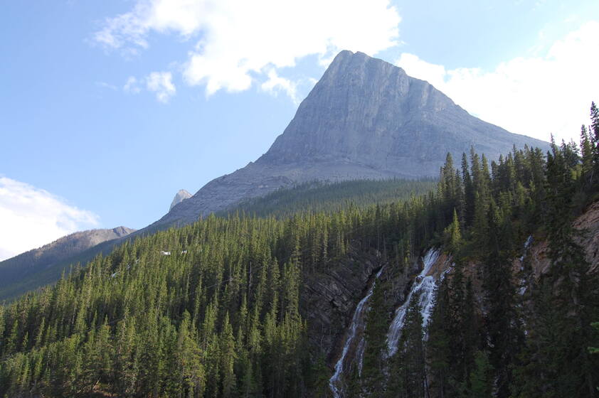 A majestic mountain rises against a clear blue sky, surrounded by lush green pine trees. A waterfall cascades down the rocky slope, adding to the scenic beauty of the landscape.