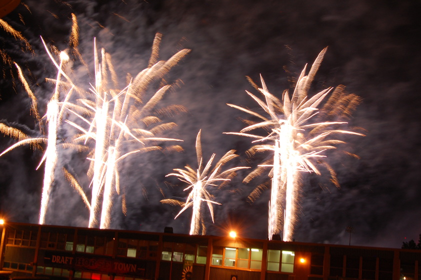 A white fireworks display erupts in a black night sky, with the silhouette of a building in the foreground.