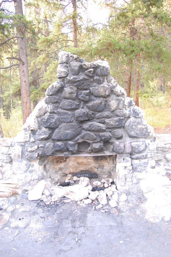 A stone fireplace surrounded by trees in a forest, with ashes and burnt wood in the fire pit.