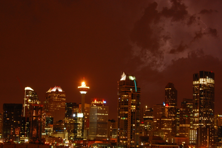 City skyline at night with illuminated skyscrapers and cloudy sky, with lightning in the distance.