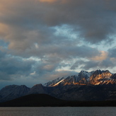 A peaceful scene of a lake at the foot of a rugged mountain range, with clouds drifting across the sky.
