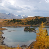 A scenic view of a lake nestled in the mountains with colorful trees in the foreground on a cloudy day.