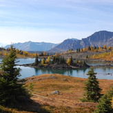 A serene alpine landscape with a clear blue lake surrounded by rugged mountains and autumn-colored trees.