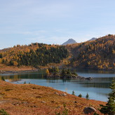 Autumn landscape with a tranquil lake surrounded by yellow and green trees, rocky mountains in the background, and a clear blue sky above.