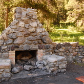 A rustic stone fireplace and seating area in a forest clearing, surrounded by trees and with remnants of a recent fire in the fireplace.