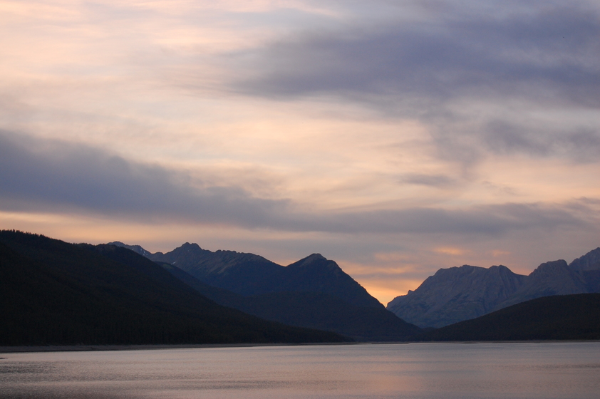A serene lake nestled amidst towering mountains at dusk, with pastel-colored clouds filling the sky.