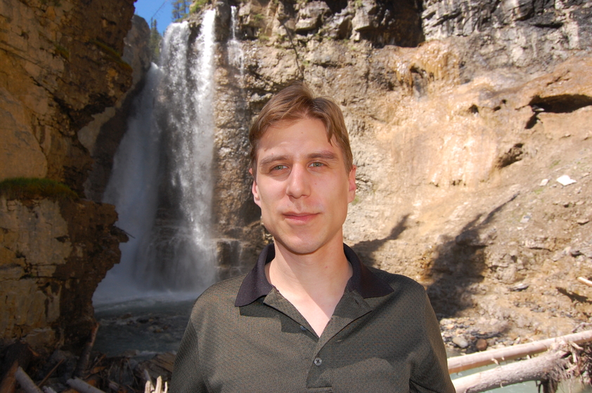 A man standing in front of a waterfall in a rocky outdoor environment.