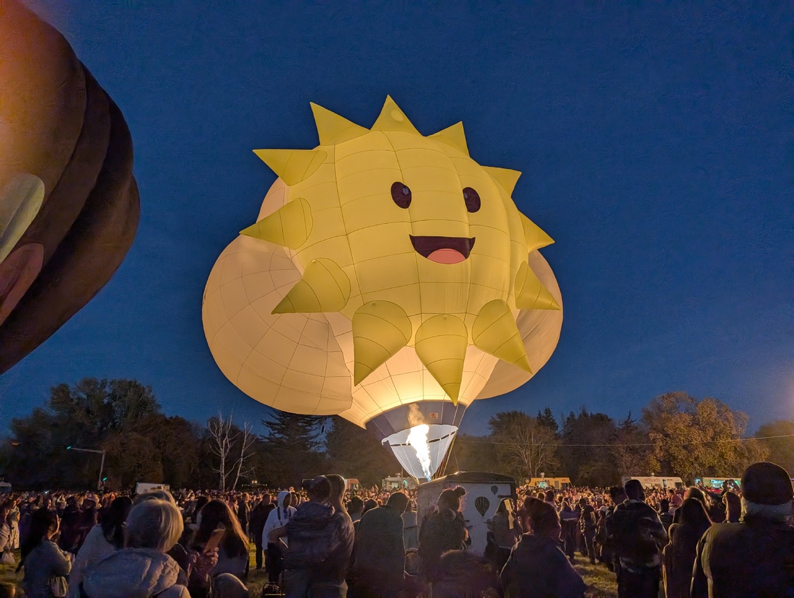 A cheerful hot air balloon with a bright yellow sun design and a smiling face, lit up against a twilight sky.