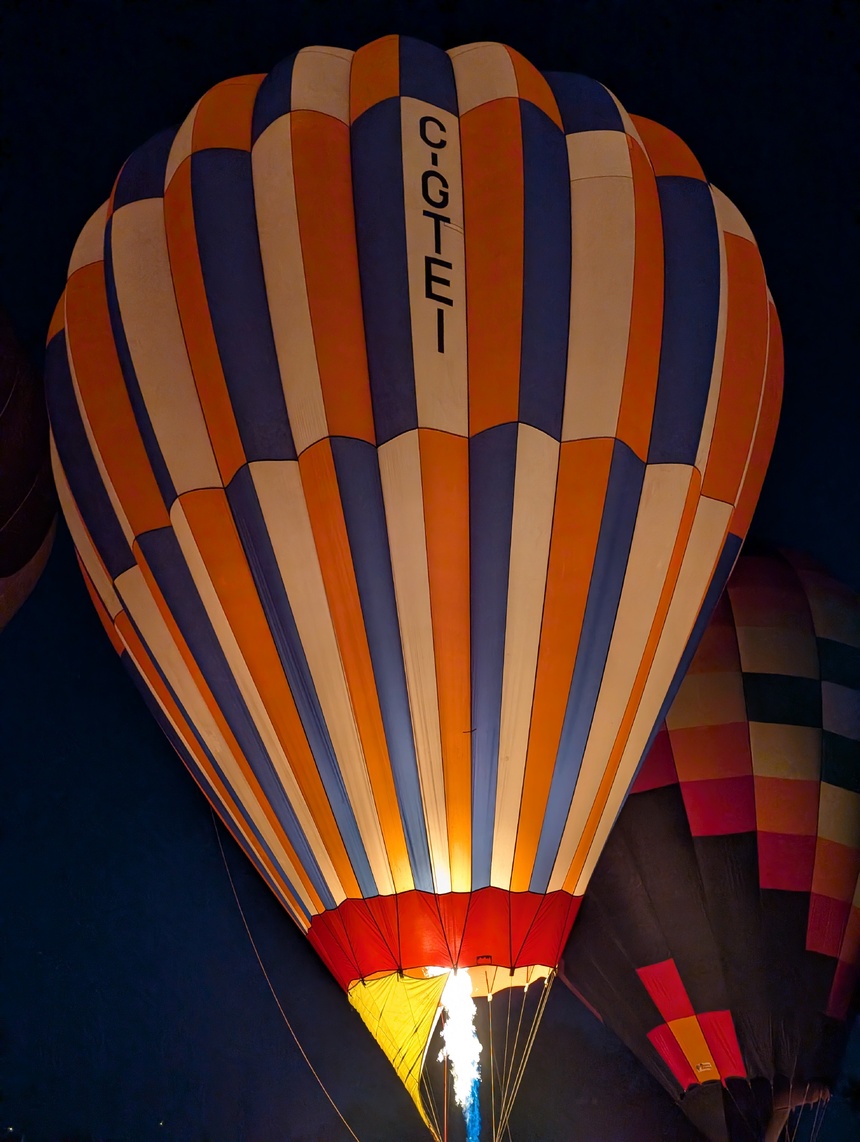 A hot air balloon with blue, orange, and white vertical stripes inflates against the night sky. The balloon's burner illuminates the interior with a warm glow, highlighting the letters "C-GTEI" vertically oriented on the balloon's surface.