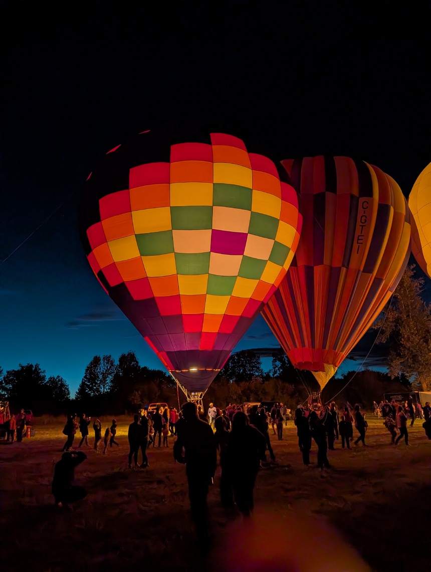 A colorful hot air balloon illuminated from within, glowing against a dark sky. It is decorated with a patchwork pattern of bright red, orange, yellow, green, and purple squares.