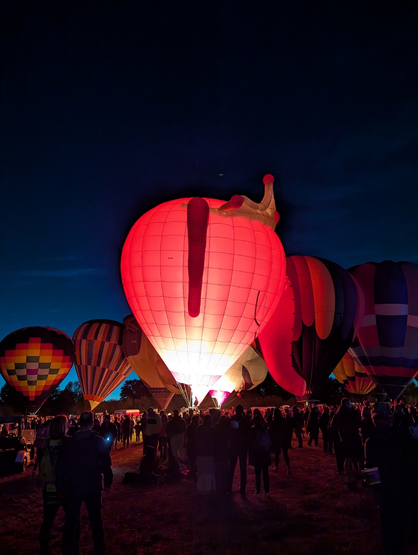 Several hot air balloons lit up against a dark blue sky. The balloon in the foreground is pink with a gold crown on top.