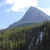 A majestic mountain rises against a clear blue sky, surrounded by lush green pine trees. A waterfall cascades down the rocky slope, adding to the scenic beauty of the landscape.