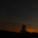 A silhouette of a person standing outdoors at night, with a starry sky and faint clouds in the background.