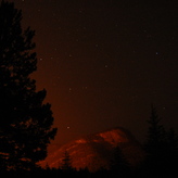 A dark night sky with silhouettes of trees in the foreground and a mountain illuminated by orange light in the background. Stars are visible scattered across the sky.