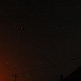 Night sky with stars visible against a dark background, with faint orange glow from city lights at the bottom.