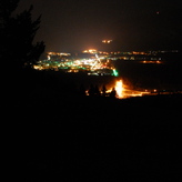 A nighttime view of Banff from a hilltop, with illuminated streets and buildings creating a glowing effect against the dark sky. Trees in the foreground frame the scene.