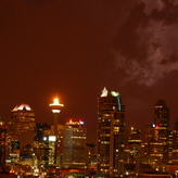 City skyline at night with illuminated skyscrapers and cloudy sky, with lightning in the distance.