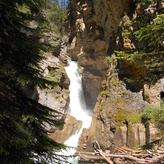 A rushing waterfall surrounded by rocky cliffs and trees in a scenic natural landscape. Fallen logs are strewn across the base of the waterfall.