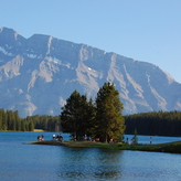 A serene lake surrounded by tall trees with a small peninsula in the center hosting a group of people enjoying outdoor activities, backed by majestic mountains.