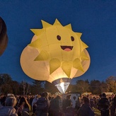 A cheerful hot air balloon with a bright yellow sun design and a smiling face, lit up against a twilight sky.