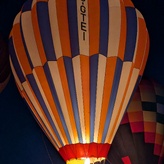 A hot air balloon with blue, orange, and white vertical stripes inflates against the night sky. The balloon's burner illuminates the interior with a warm glow, highlighting the letters "C-GTEI" vertically oriented on the balloon's surface.