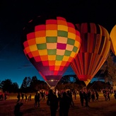 A colorful hot air balloon illuminated from within, glowing against a dark sky. It is decorated with a patchwork pattern of bright red, orange, yellow, green, and purple squares.