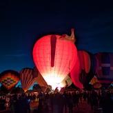 Several hot air balloons lit up against a dark blue sky. The balloon in the foreground is pink with a gold crown on top.